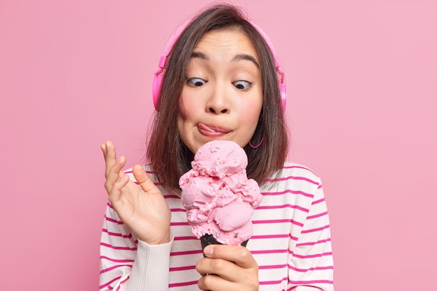 Mujer mira apetitoso helado lame labios come delicioso postre helado escucha música a través de auriculares vestida con jersey de rayas