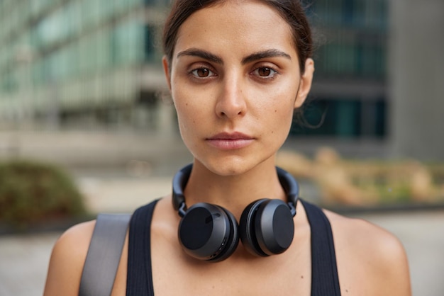 mujer mira atentamente a la cámara lo motiva a practicar deporte usa audífonos inalámbricos alrededor del cuello vestida con ropa deportiva va a tener poses de entrenamiento físico al aire libre