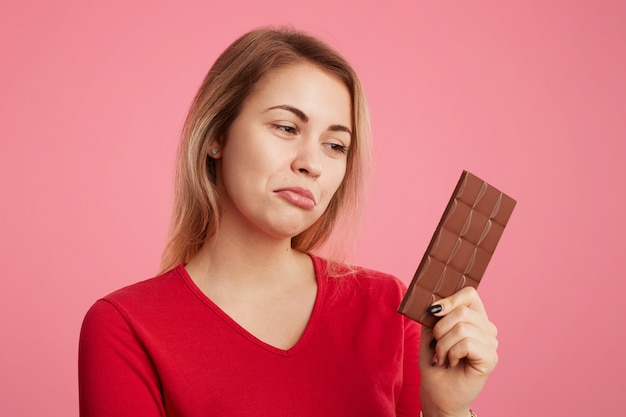 Foto gratuita la mujer mira con expresión de descontento la dulce barra de chocolate, sigue la dieta, no puede comerla para ser delgada y deportiva