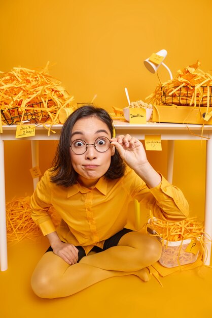 La mujer mira fijamente, no puedo creer que sus ojos usen gafas redondas, ropa elegante, posa en el gabinete en el espacio de coworking en amarillo.