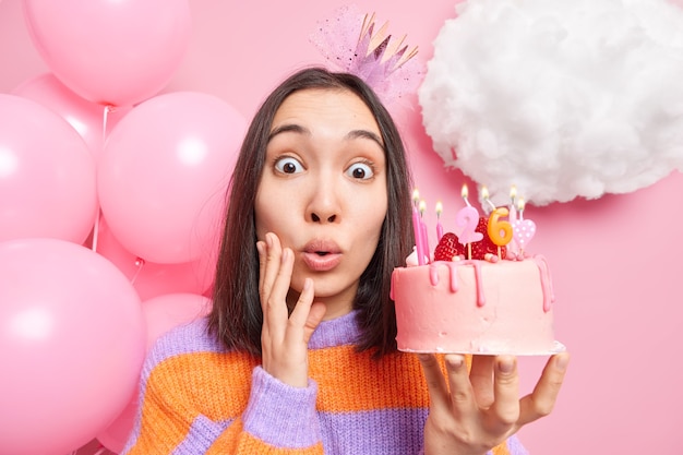 Foto gratuita mujer mira fijamente sorprendida a la cámara sostiene un delicioso pastel de cumpleaños sabroso lleva corona y poses de puente rayado