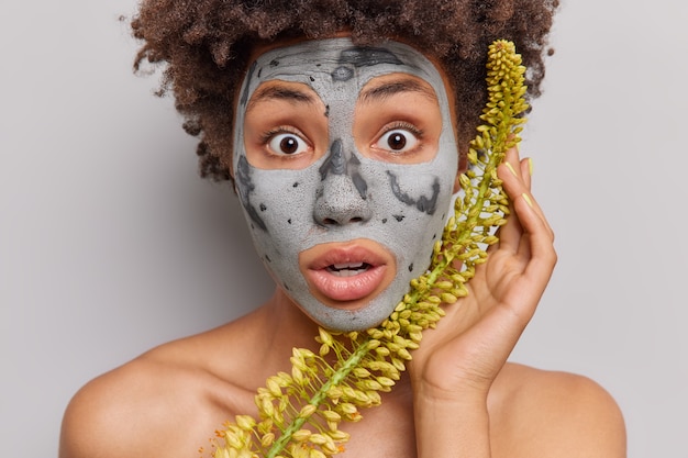 Foto gratuita mujer mira impresionada a la cámara aplica mascarilla de arcilla a base de hierbas no puedo creer que sus ojos estén en topless interior en gris