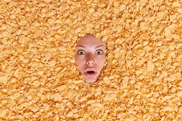 Foto gratuita mujer mira impresionada rodeada de cereales secos tiene mirada atenta se siente asustada come comida sana quiere tener un delicioso refrigerio para el desayuno
