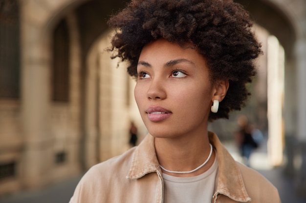 Mujer mira hacia otro lado pensativamente viste chaqueta beige aretes mira en algún lugar plantea paseos en la ciudad piensa en el futuro