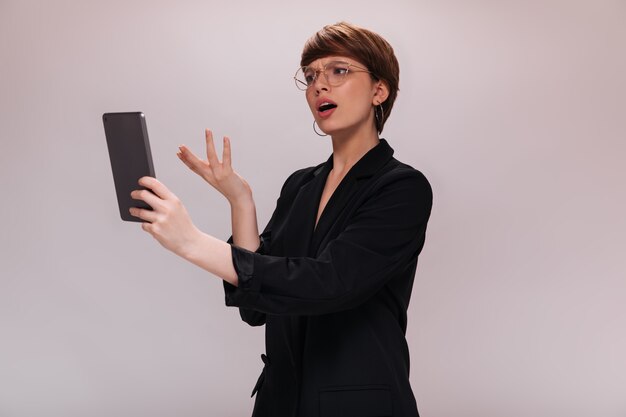 La mujer mira a la pantalla de la tableta con malentendidos. Señora de negocios en poses de chaqueta negra sobre fondo blanco aislado