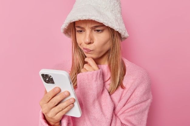 La mujer mira la pantalla del teléfono inteligente lee la notificación recibe un mensaje de una persona desconocida usa un sombrero blanco y un puente aislado en rosa tiene una expresión dudosa