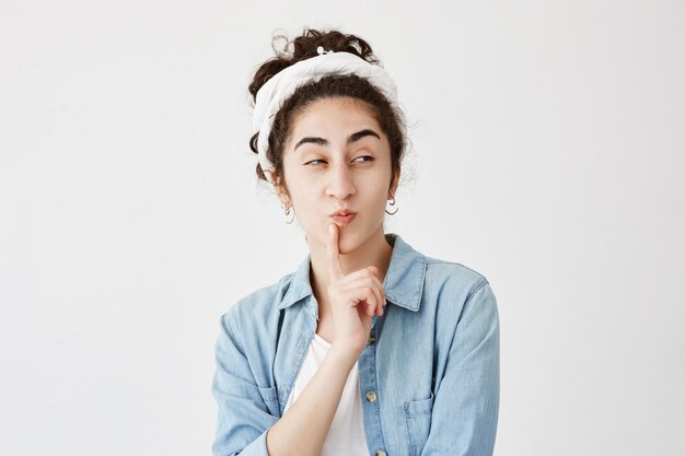 La mujer mira pensativamente a un lado con cabello oscuro y ondulado, trata de tomar una decisión, mantiene el dedo en los labios, posa contra la pared con espacio para copiar texto promocional. Concepto de personas y lenguaje corporal