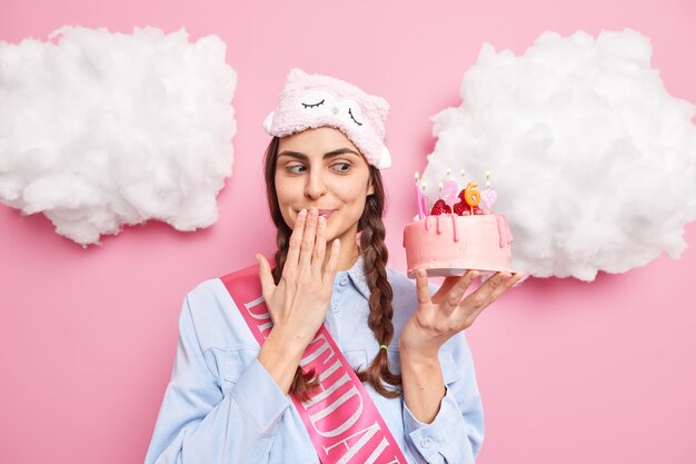 La mujer mira con tentación a un delicioso pastel contra la boca espera a los invitados en la fiesta de cumpleaños viste una camisa sleepmask aislada en rosa