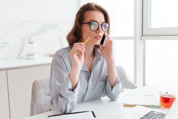 Mujer con mirada pensativa sosteniendo un lápiz y hablando por teléfono inteligente mientras se encuentra en el lugar de trabajo en la sala blanca