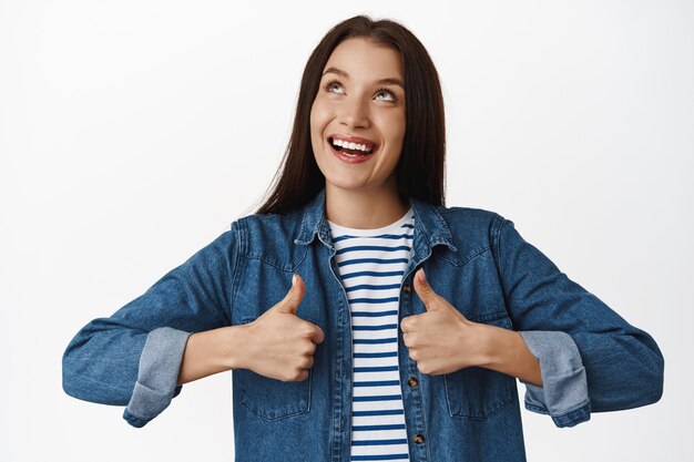 mujer, mirando hacia arriba y sonriendo, mostrando los pulgares hacia arriba, como aprobar, recomendar producto en la parte superior, satisfecha con los precios de venta, de pie en blanco.