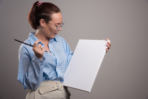 Mujer mirando lienzo vacío y pincel sobre fondo gris