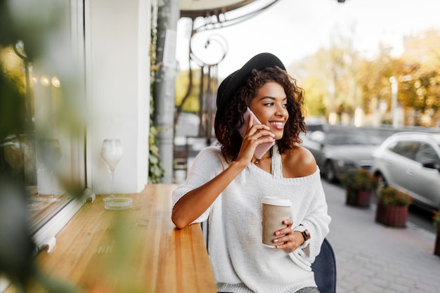 Mujer mixta con peinado afro hablando por teléfono móvil y sonriendo en segundo plano urbano. Chica negra con ropa casual. Sosteniendo la taza de café. Sombrero negro.