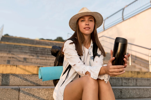 Mujer con mochila y sombrero sosteniendo termo mientras viaja