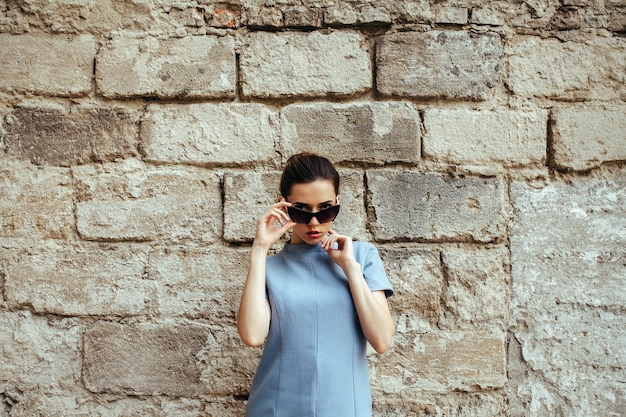 Mujer de moda atractiva en vestido azul con gafas de sol posando junto a la pared blanca
