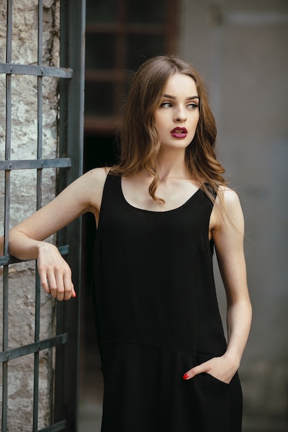 Mujer de moda atractiva en vestido negro posando junto a la pared blanca