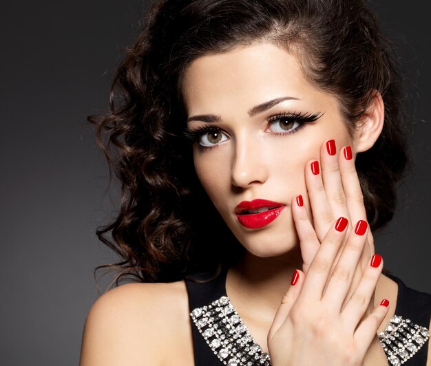 Mujer de moda de belleza con uñas rojas, labios y maquillaje de ojos dorados - en pared negra