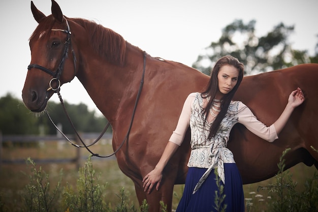 Mujer de moda con cabello castaño largo posando con caballo marrón.