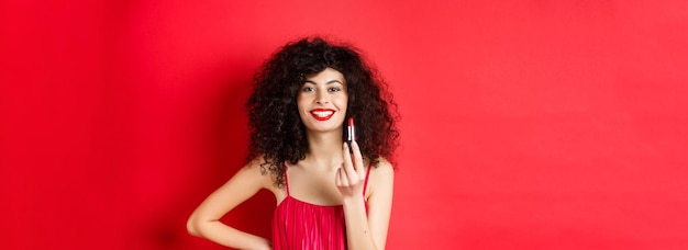 Foto gratuita mujer de moda con cabello rizado que muestra lápiz labial rojo y sonriente recomienda pie cosmético en wh