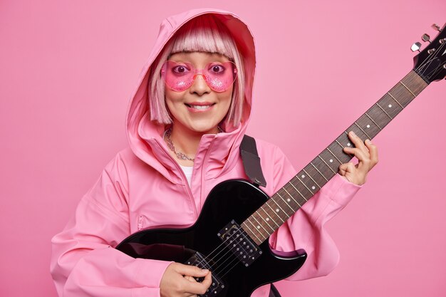 Mujer de moda con cabello rosado finge actuar en el escenario toca música rock and roll usa gafas de sol en forma de corazón y anorak posa en el interior contra la pared rosada. Solista talentosa