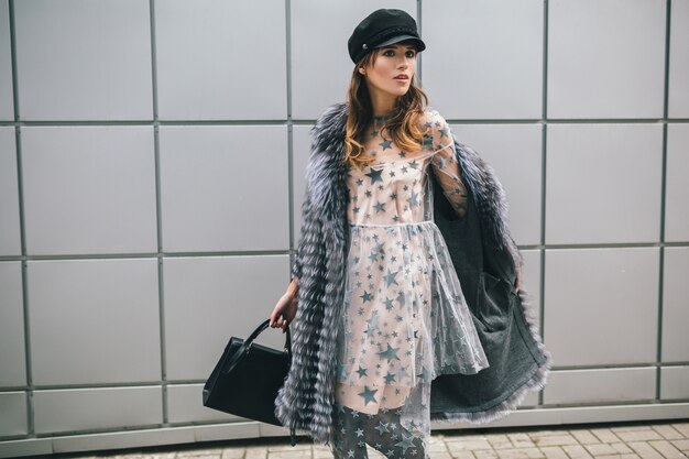 Mujer de moda caminando en la ciudad en abrigo de piel cálido y vestido de fiesta, accesorios de temporada de invierno