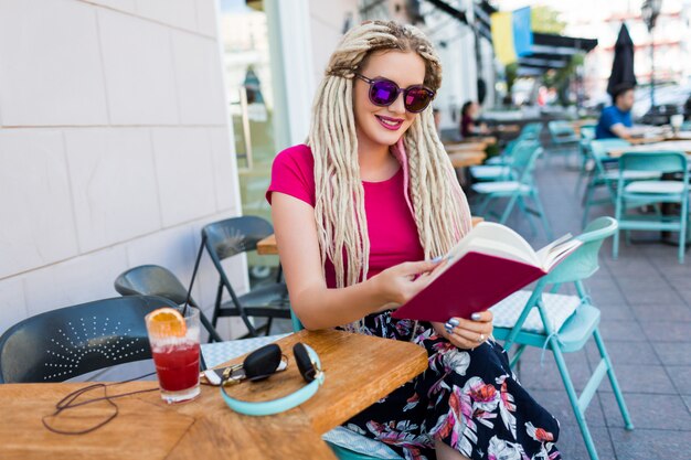 Mujer de moda elegante con rastas blancas en gafas de sol sosteniendo el cuaderno y pasando su tiempo libre en un restaurante moderno. Batido fresco y auriculares en la mesa.