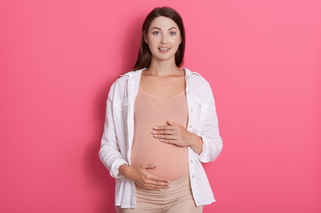 Mujer de moda embarazada atractiva joven con ropa casual, mirando directamente a la cámara, teniendo emociones positivas. cámara, tocando su vientre con ambas manos contra el espacio rosa.