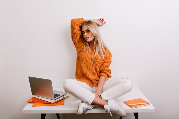 Mujer de moda feliz en pantalones blancos de moda jugando en la oficina, sentada con las piernas cruzadas en la mesa cerca de la computadora