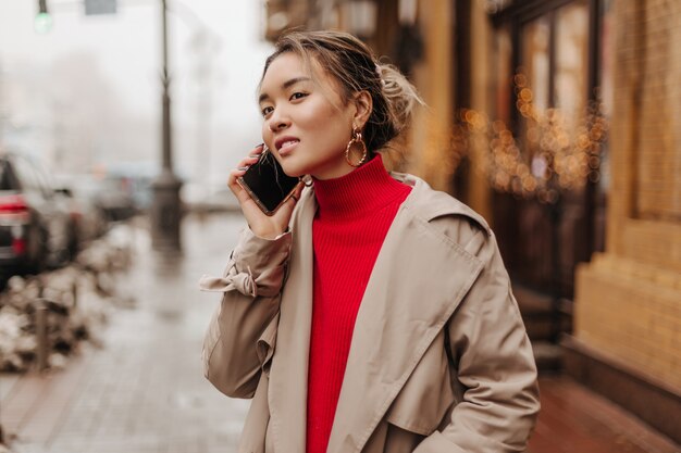 Mujer de moda con gabardina ligera y suéter brillante hablando por teléfono de muy buen humor y caminando por la ciudad