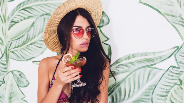 Mujer de moda en gafas de sol de corazón rosa brillante bebe cóctel de fresa fresca en el bar de la playa.