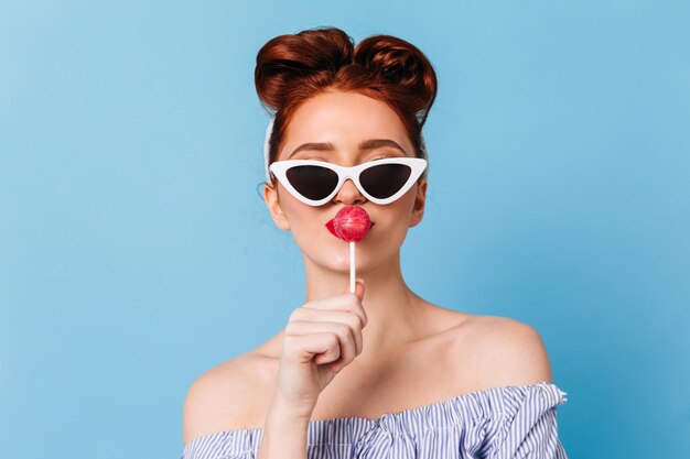 Mujer de moda en gafas de sol negras lamiendo piruleta Vista frontal de dama pinup con caramelo aislado sobre fondo azul