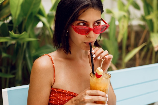 Mujer de moda en gafas de sol retro relajantes en club de playa tropical en elegante top rojo y pantalones cortos de color naranja. Beber limonada sabrosa.