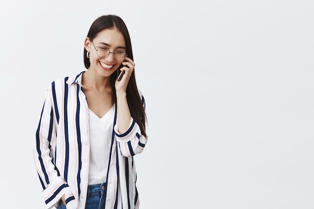 Mujer de moda joven alegre y exitosa hablando por teléfono, disfrutando de la conversación. Mujer relajada despreocupada en blusa a rayas y gafas, mirando hacia abajo con linda sonrisa, sosteniendo el teléfono inteligente