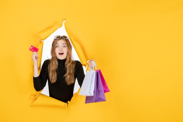 Mujer de moda joven sosteniendo bolsas de la compra y tarjeta de crédito a través del agujero de papel rasgado en la pared