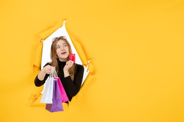 Mujer de moda joven sosteniendo bolsas de la compra y tarjeta de crédito a través del agujero de papel rasgado en la pared