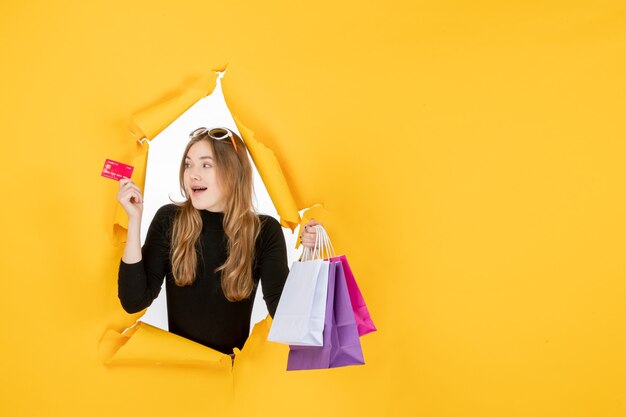 Mujer de moda joven sosteniendo bolsas de la compra y tarjeta de crédito a través del agujero de papel rasgado en la pared