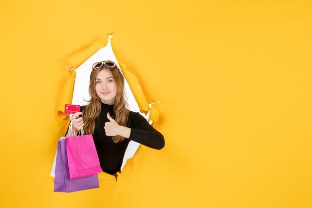 Mujer de moda joven sosteniendo bolsas de la compra y tarjeta de crédito a través del agujero de papel rasgado en la pared