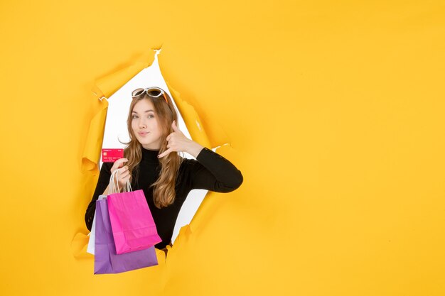 Mujer de moda joven sosteniendo bolsas de la compra y tarjeta de crédito a través del agujero de papel rasgado en la pared