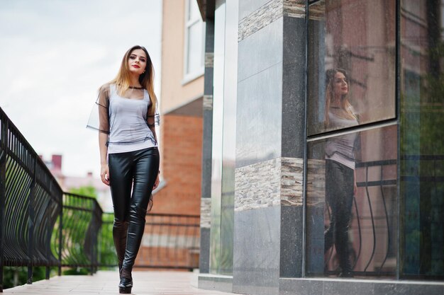 Mujer de moda mira camisa blanca ropa transparente negra pantalones de cuero posando en la calle contra grandes ventanales del edificio Concepto de chica de moda