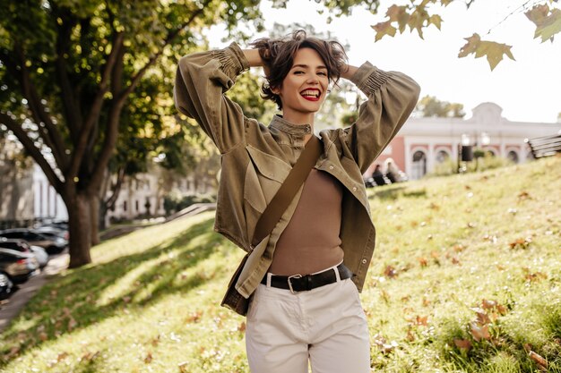 Mujer de moda con peinado morena con bolso sonriendo al aire libre. Una mujer fresca con chaqueta verde oliva y pantalones blancos se ríe afuera.