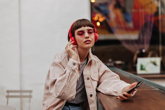 Mujer de moda con el pelo corto en auriculares escuchando música al aire libre Hermosa chica en chaqueta de mezclilla fresca y gafas brillantes sosteniendo el teléfono en la cafetería