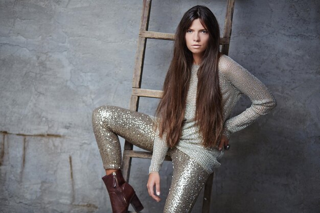 Mujer de moda con el pelo largo y castaño con un traje brillante posando sobre una pared gris con una escalera de madera.