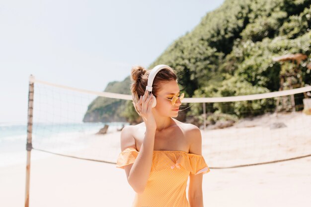 Mujer de moda en ropa amarilla posando delante de la red de voleibol. Foto al aire libre de chica caucásica bronceada en auriculares escuchando música en la playa