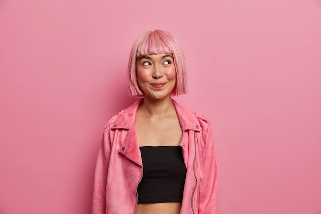 Foto gratuita la mujer de moda tiene el cabello rosado y flecos, se ve con una expresión feliz y soñadora, mira hacia arriba, viste un top negro con una elegante chaqueta rosada. emociones agradables, concepto de estilo