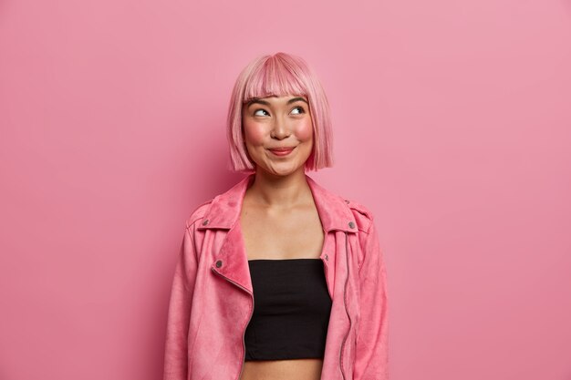 La mujer de moda tiene el cabello rosado y flecos, se ve con una expresión feliz y soñadora, mira hacia arriba, viste un top negro con una elegante chaqueta rosada. Emociones agradables, concepto de estilo