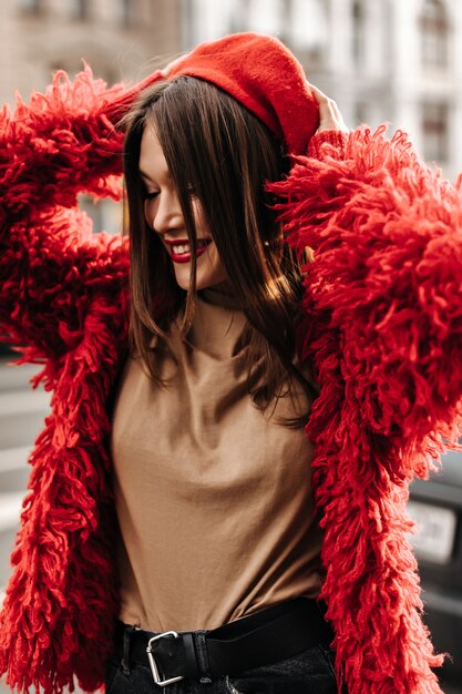 Mujer de moda en top beige y abrigo rojo se pone boina al estilo francés, caminando por la ciudad.