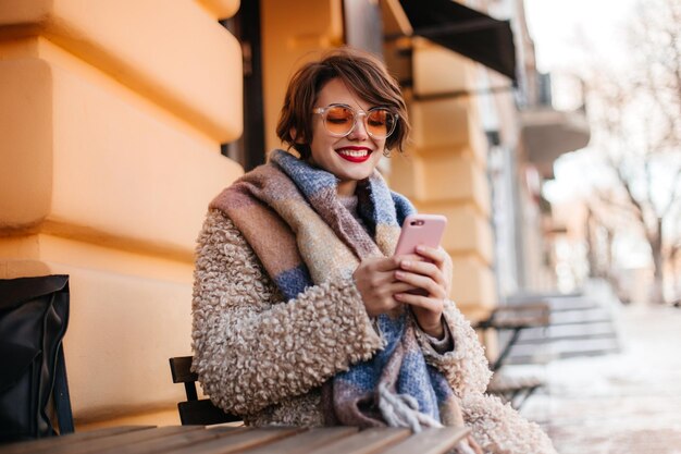 Mujer de moda usando un teléfono inteligente en un día frío Toma al aire libre de una chica guapa sonriente en abrigo con bufanda