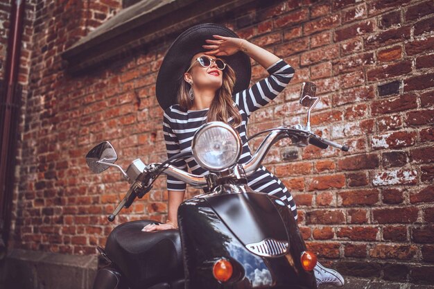 Mujer de moda vestida con rayas posando cerca de scooter sobre la pared de ladrillos rojos.