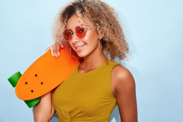 Mujer modelo sonriente en jeans cortos de moda de verano y gafas de sol posando con skate