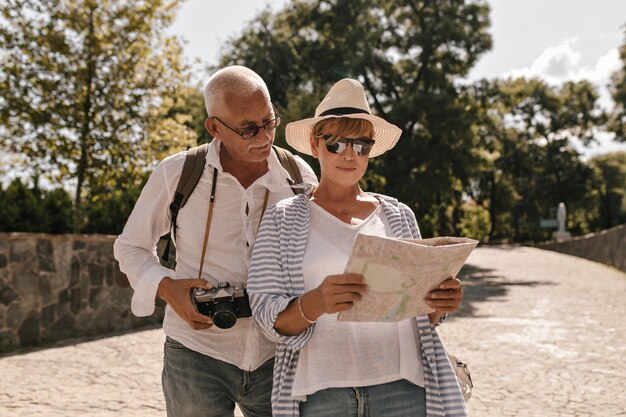 Mujer moderna con camiseta blanca, blusa a rayas, sombrero y gafas mirando el mapa y caminando con un hombre de pelo gris con camisa y cámara al aire libre
