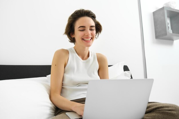 Foto gratuita mujer moderna descansando en casa sentada en la cama con una computadora portátil mirando la pantalla de la computadora riendo y